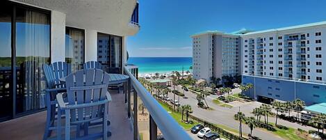 A balcony with chairs, a view of the ocean, and a parking lot with cars.