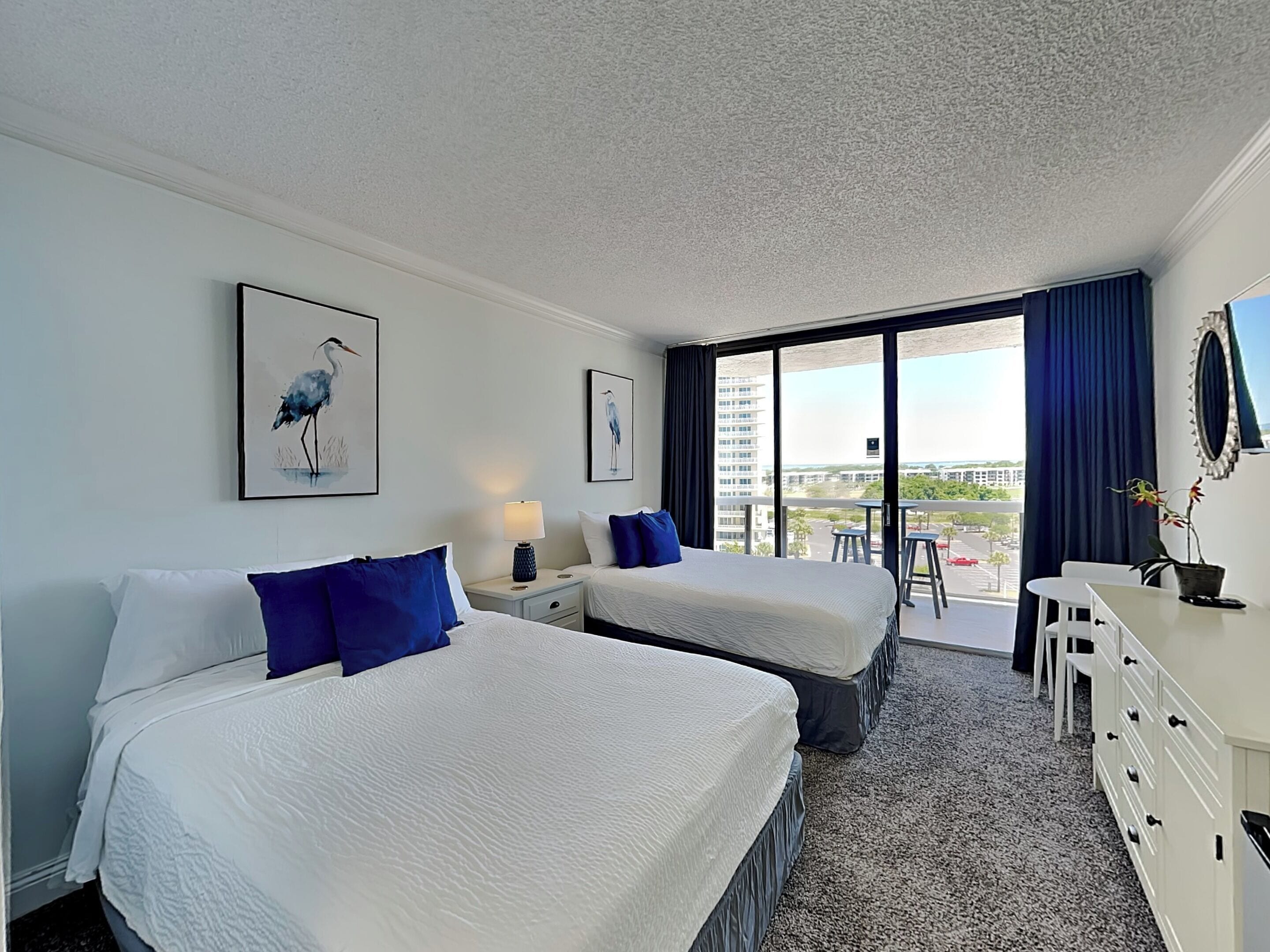 A hotel room with two beds, a desk, a chair, a lamp, and a large window with a view of buildings.