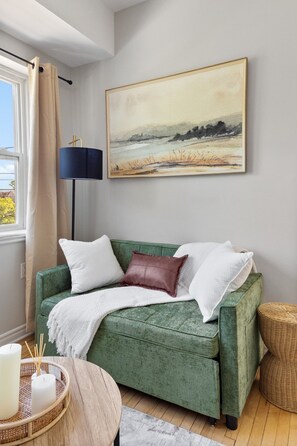 Ein grünes Sofa mit weißen und braunen Kissen, eine Lampe, ein Gemälde und ein Korbtisch.