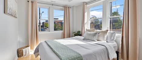 A bedroom with a large bed, two windows, and a view of buildings outside.