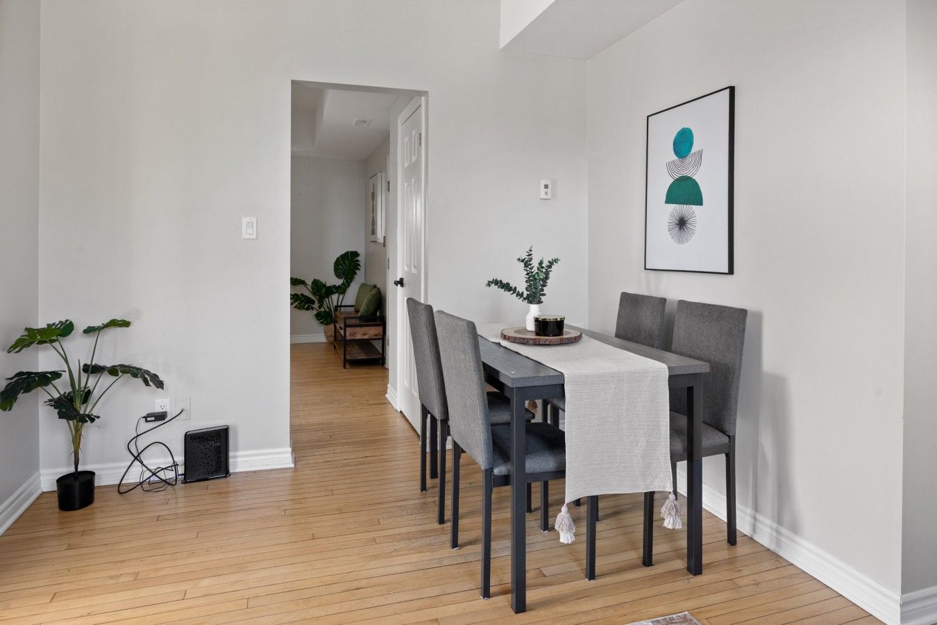 Uma área de refeições com piso de madeira, uma mesa com toalha branca e cadeiras cinzas. Há uma obra de arte emoldurada na parede e um vaso de planta sobre a mesa.