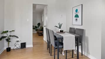 A dining area with a wooden floor, a table with a white tablecloth, and grey chairs. There is a framed artwork on the wall and a potted plant on the table.