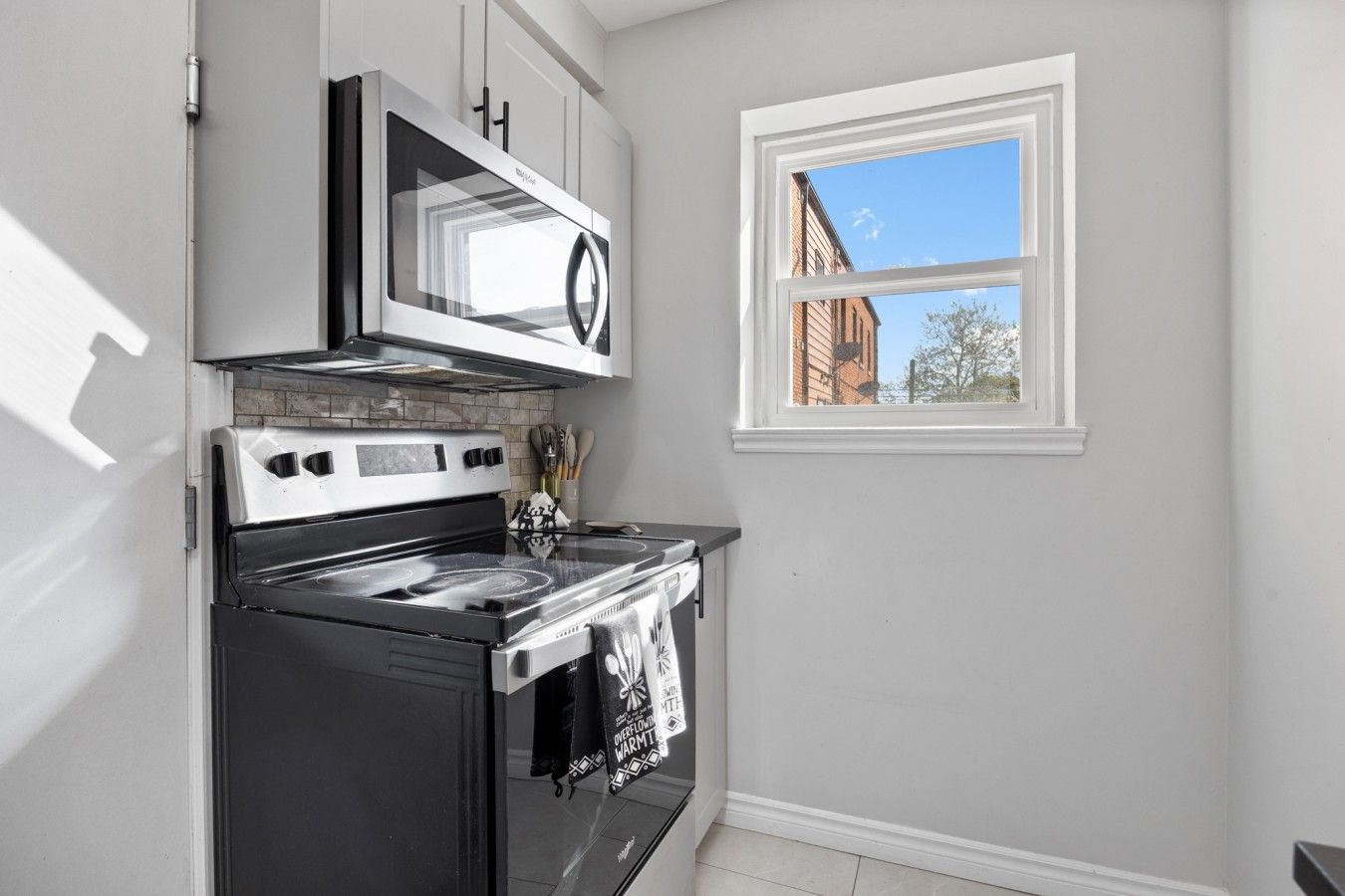 A kitchen with a stove, microwave, and oven, a window with a view of buildings, and a tiled backsplash.