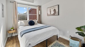 Quarto com cama, janela, mesinha de apoio, planta e quadro pendurado na parede.