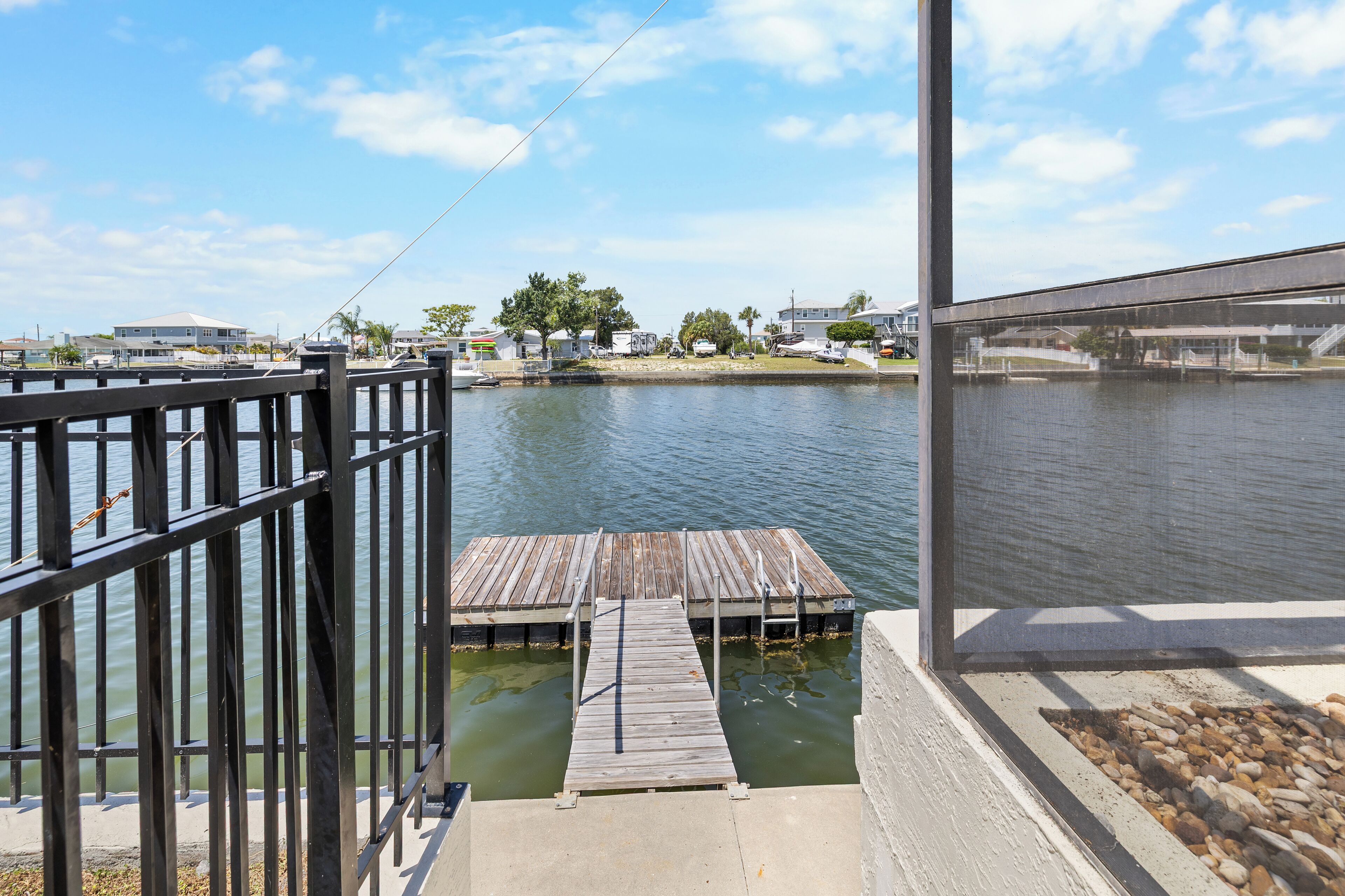 A waterfront dock with a wooden walkway leading to a pier, surrounded by calm water and distant houses.