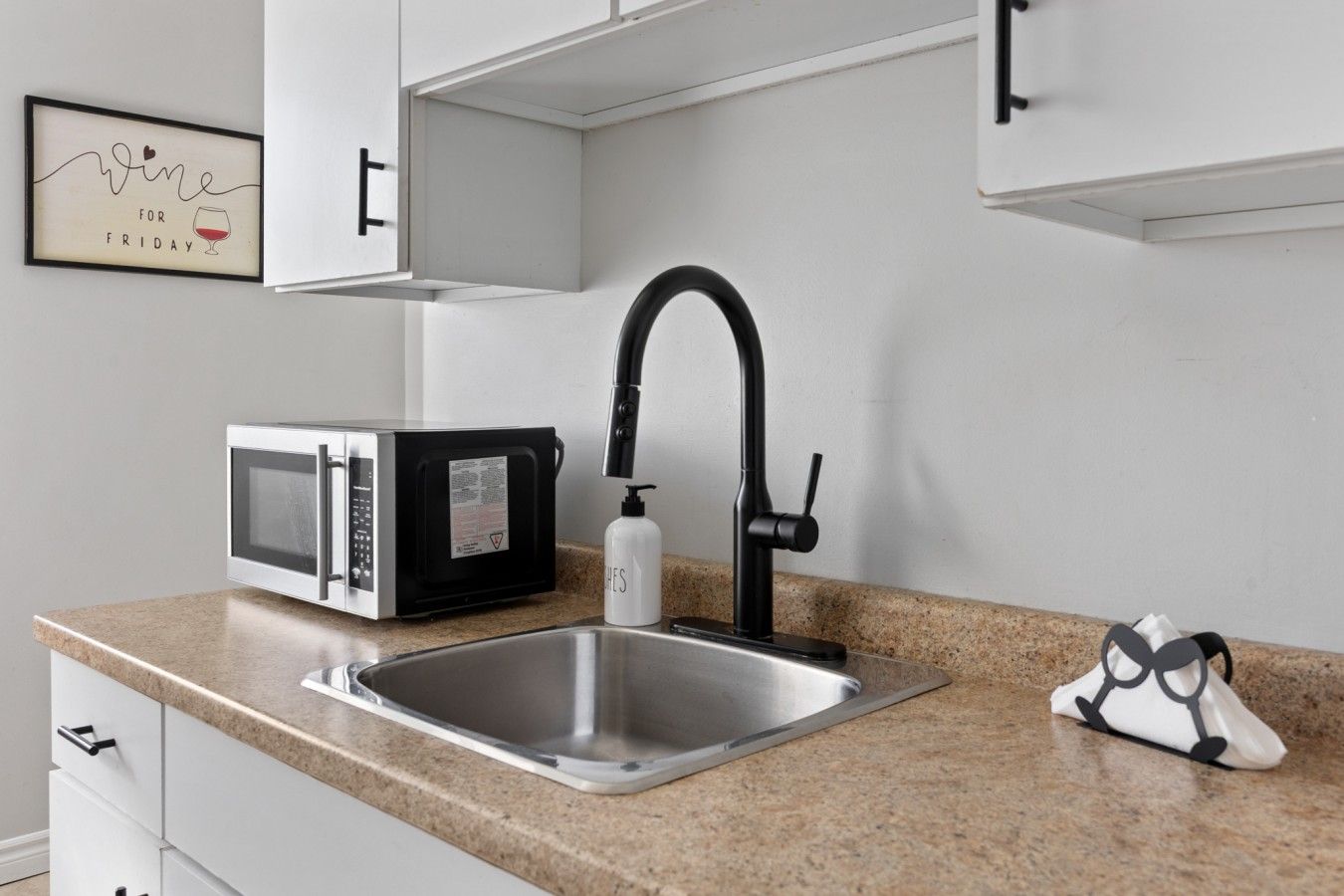 A kitchen with a stainless steel sink, a black faucet, a microwave, and a framed sign on the wall.