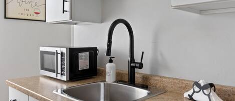 Eine Küche mit einem Edelstahl-Spülbecken, einem schwarzen Wasserhahn, einer Mikrowelle und einem gerahmten Schild an der Wand.