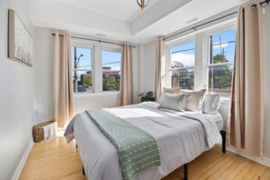 A bedroom with a large bed, two windows, and a view of buildings outside. - Walkerville Windsor | 3BR | 2 Private Apartments | Sleeps 6 (Windsor)