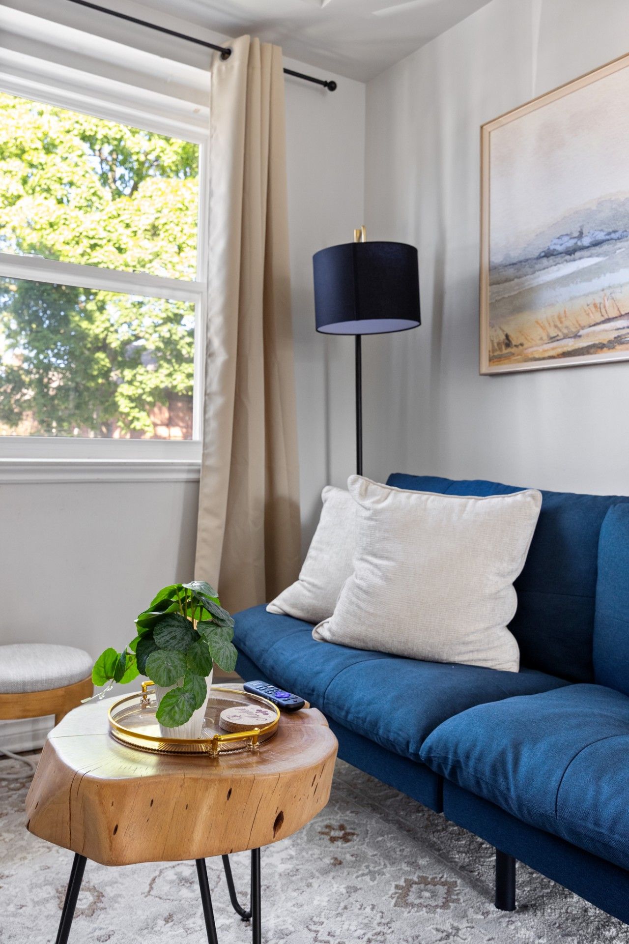 A living room with a blue sofa, a round wooden coffee table, a floor lamp, a painting on the wall, and a window with curtains.