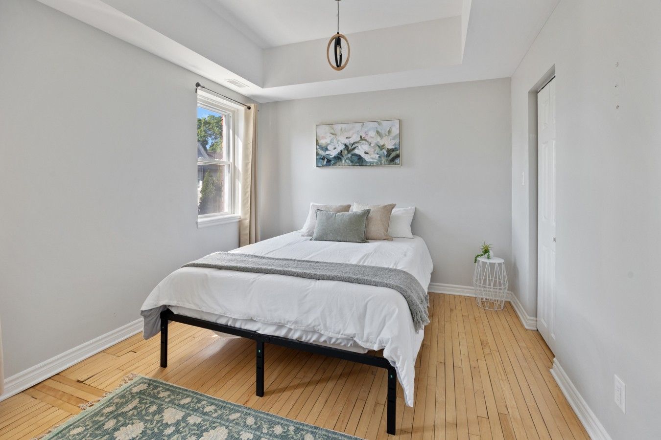 A bedroom with a bed, a window, a painting, and a small white table.