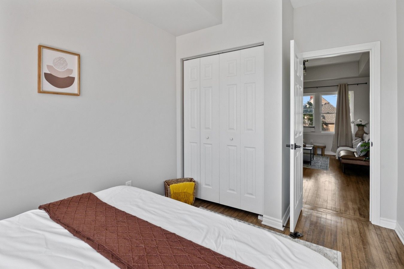 A bedroom with a bed, a white door, a wicker chair, and a hallway leading to a living area.