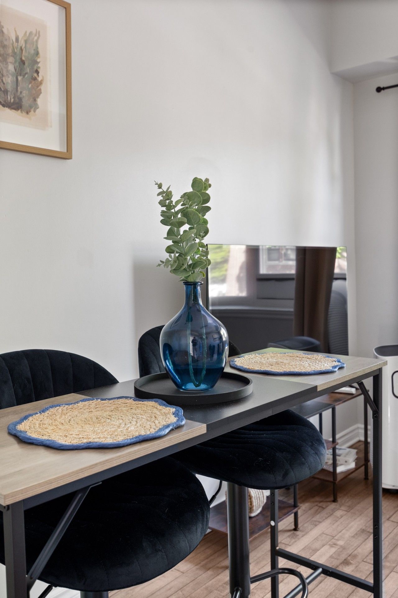 A modern dining area with a wooden table, black chairs, a blue vase with green plants, and a woven placemat.