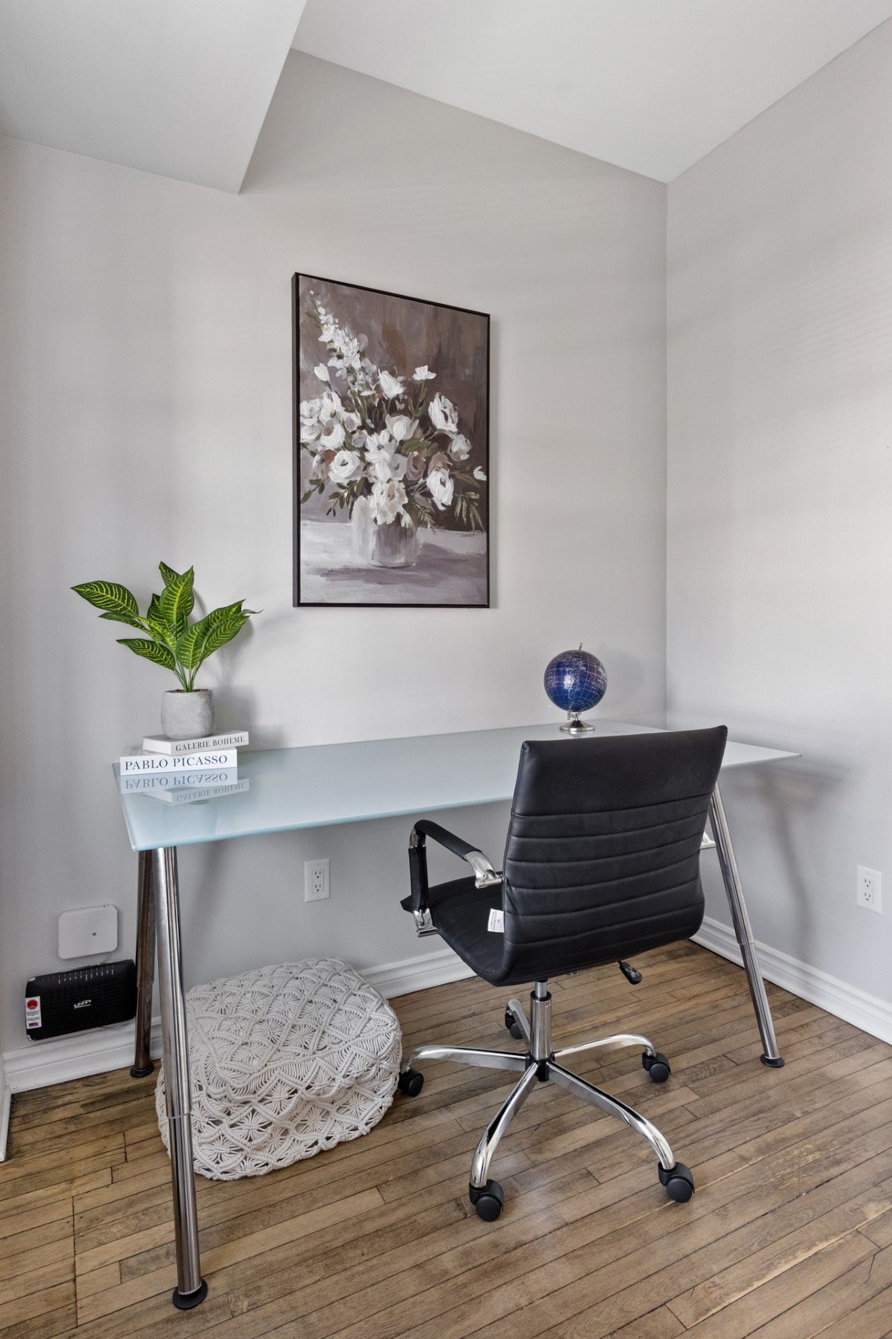 A modern office space with a glass desk, a black office chair, a potted plant, a globe, and a framed painting on the wall.