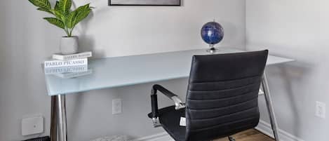A modern office space with a glass desk, a black office chair, a potted plant, a globe, and a framed painting on the wall.