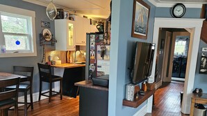 A kitchen with wooden flooring, a dining area with chairs, a TV on a stand, and a clock on the wall.