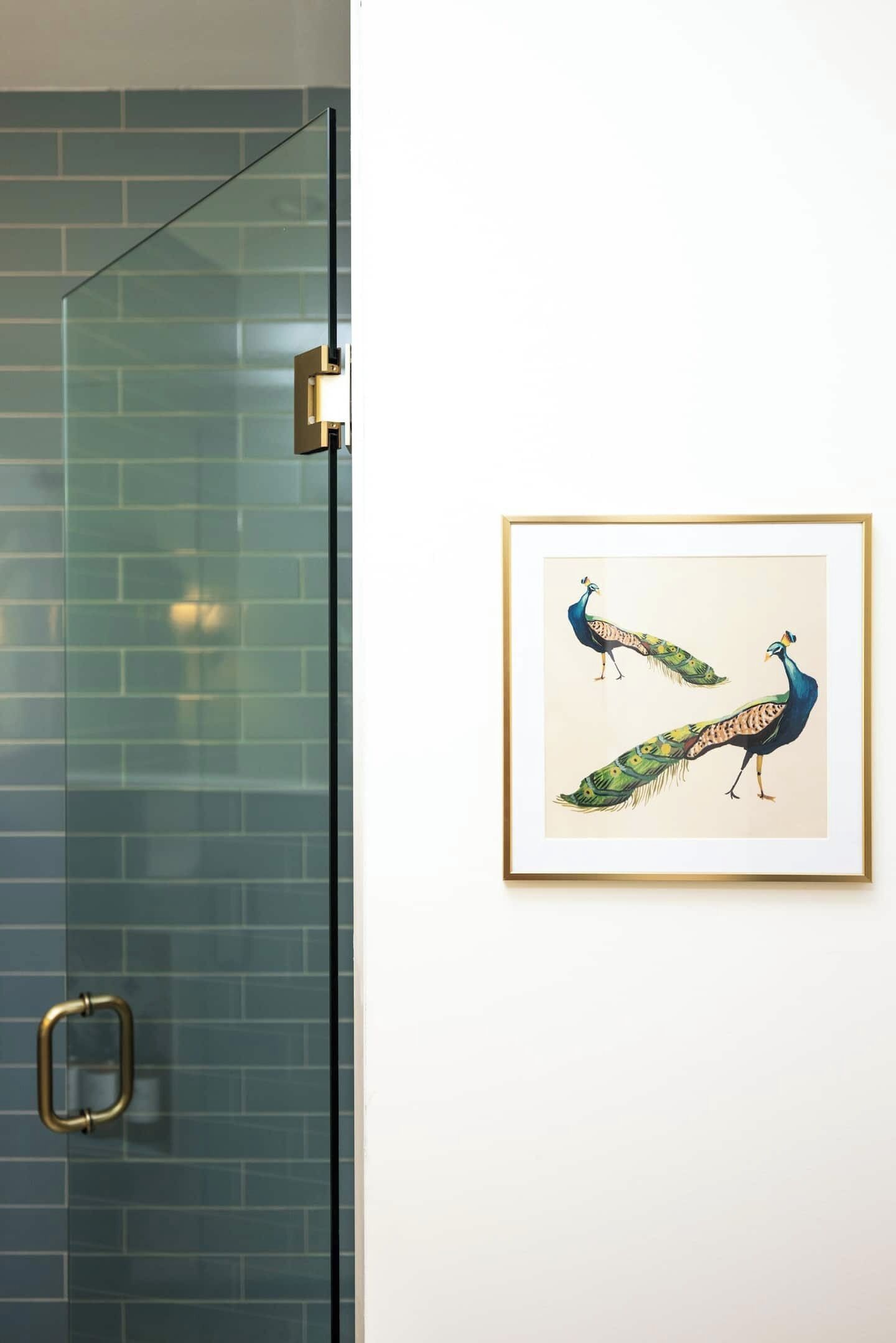 A framed peacock artwork on a white wall next to a glass shower door.
