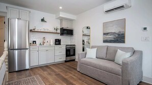 Et moderne køkken med hvide skabe, et stålkøleskab, en mikroovn og en ovn. Der er en grå sofa med puder og et tæppe på gulvet.