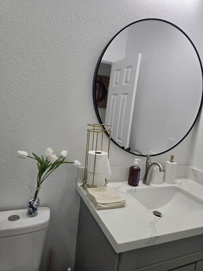 Ein Badezimmer mit einem weißen Waschbecken, einer Toilette, einem Spiegel mit schwarzem Rahmen, einer Vase mit weißen Blumen und einer Toilettenpapierrolle.