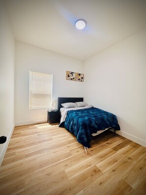 Un dormitorio con cama, mesita de noche, ventana con persianas y una lámpara en el techo.