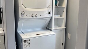 Une petite buanderie équipée d’un lave-linge et d’un sèche-linge à chargement frontal, d’une petite étagère et d’une porte donnant sur une autre pièce.