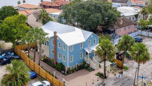 Et kystnære boligområde med et markant blå hus med ståltag, omgivet af palmer og et træhegn.
