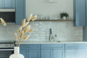 Una cocina con armarios azules, un jarrón blanco con espigas de trigo secas y un salpicadero de azulejos blancos.