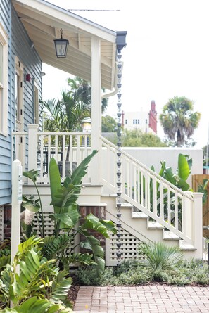 Una casa con un porche blanco, una escalera y un jardín con diversas plantas.
