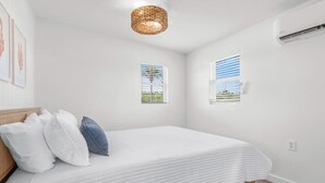Ein Schlafzimmer mit einem Bett, Kissen, einer Hängelampe, zwei Fenstern mit Jalousien und einer Klimaanlage.
