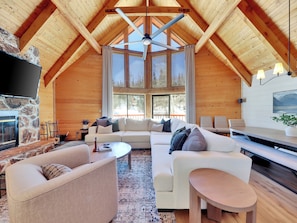 A log cabin interior with a stone fireplace, a large sofa, a television, and a dining area. - Fall in Brian Head means golden aspens, crisp mountain air & quieter trails. This 5-bedroom retreat pairs outdoor adventure with a hot tub, fire pit, chef’s kitchen, game room and restorative spaces for families, friends and multi-generational stay. (Brian Head)