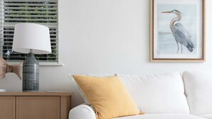 Ein Zimmer mit einem weißen Sofa, einem gelben Kissen, einer Lampe und einem Bilderrahmen mit einem Vogelbild an der Wand.