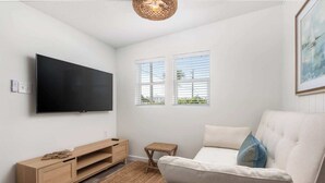 Ein modernes Wohnzimmer mit Flachbildfernseher, einer Holz-Unterhaltungsecke, einer gepolsterten Couch, einem kleinen Hocker und zwei Fenstern mit Jalousien.