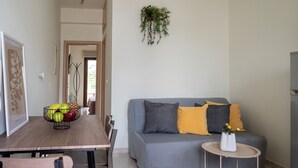 Ein Wohnzimmer mit grauem Sofa, einem kleinen runden Tisch, einem Esstisch mit Stühlen und einer hängenden Pflanze.