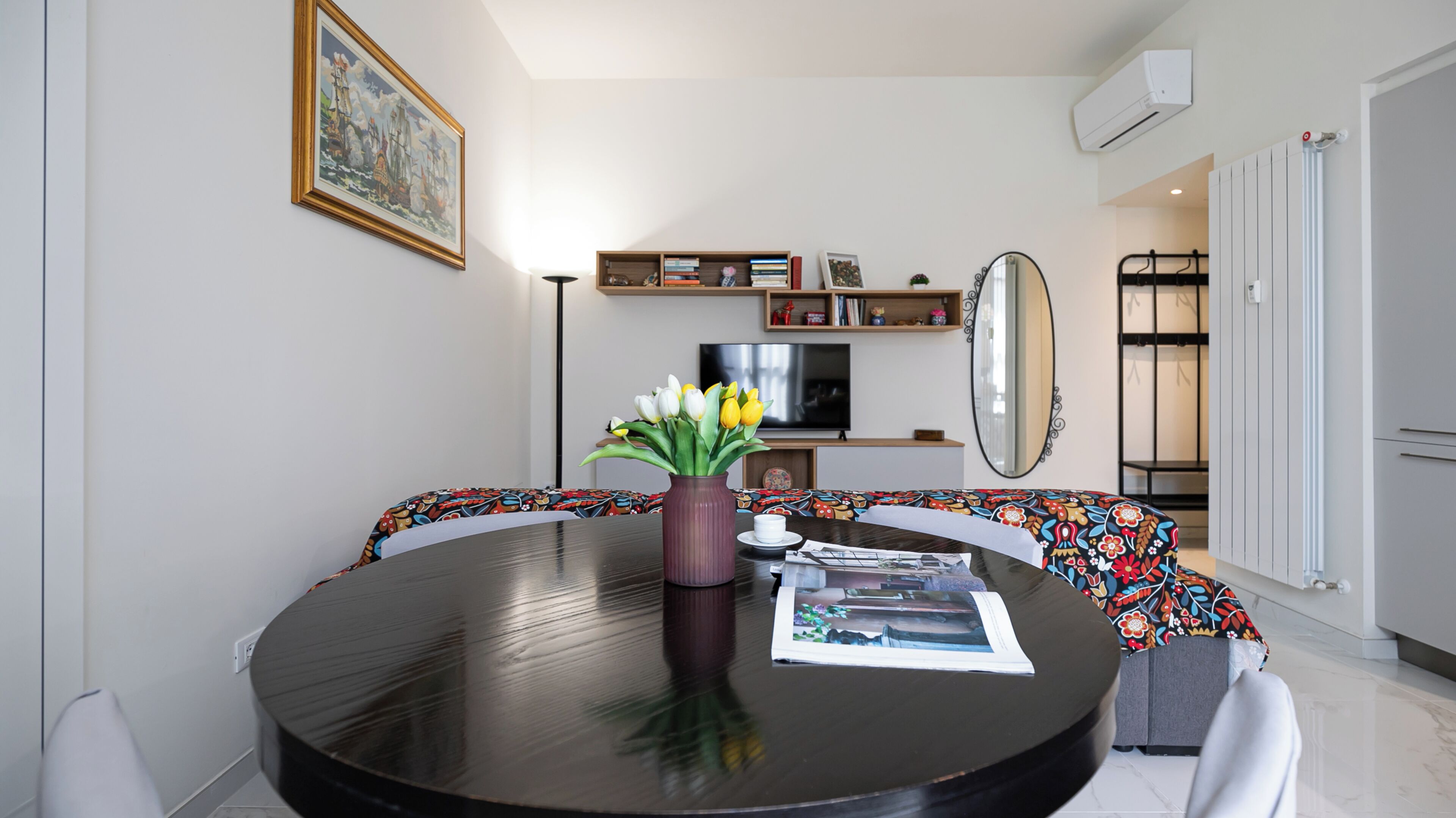 Un salon moderne avec une table à manger ronde, un vase de fleurs, un téléviseur à écran plat, une bibliothèque et un grand miroir.