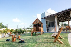 Un terrain de jeux doté de structures en bois, notamment une balançoire et une petite maison servant de cabane, situé dans un espace verdoyant avec un bâtiment en arrière-plan.