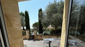 Vista a través de un ventanal hacia un patio exterior con suelo de baldosas, una mesa con sillas y una piscina al fondo.