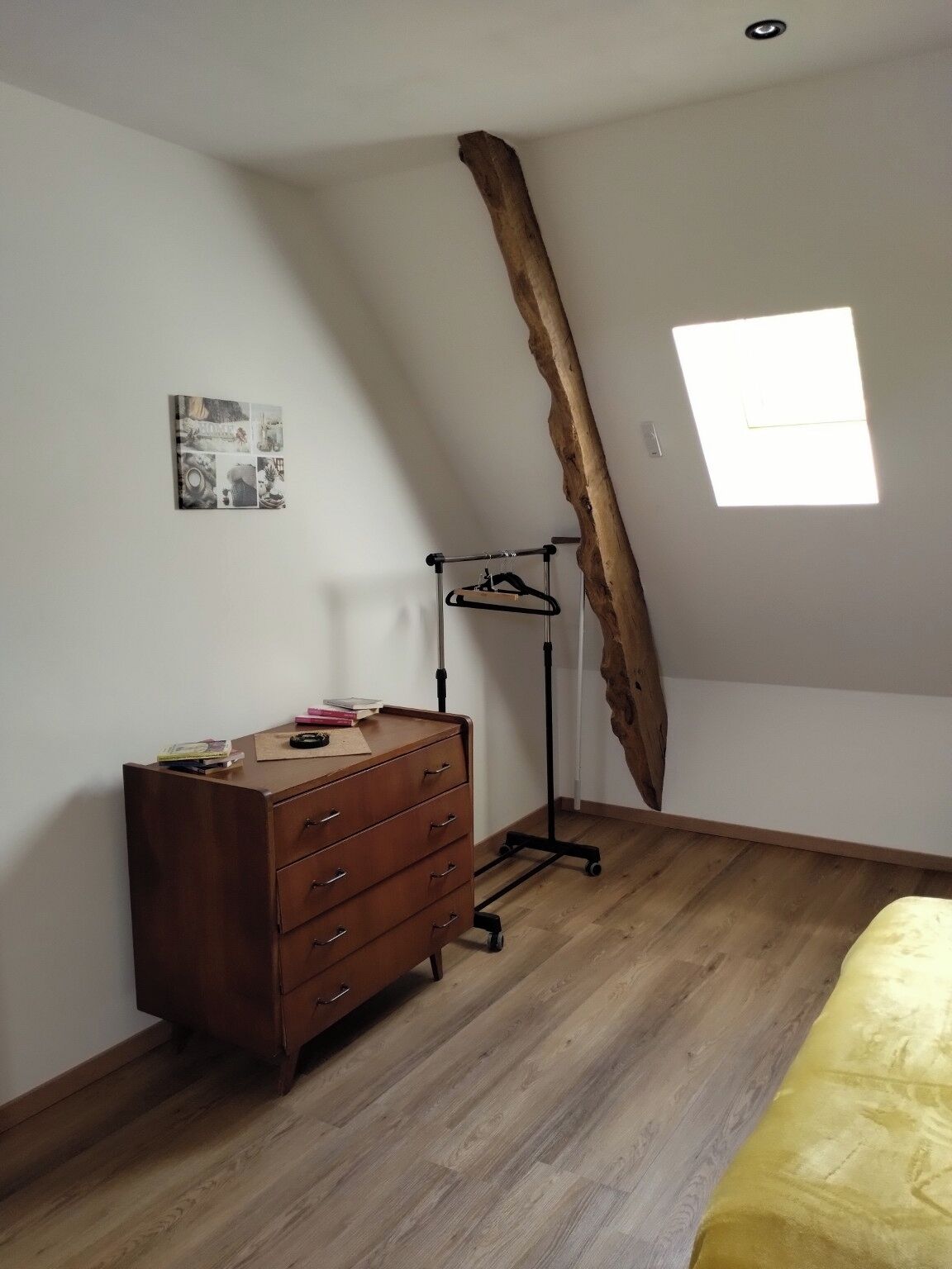 A bedroom with a wooden dresser, a hanging clothes rack, a window with natural light, and a wall-mounted picture.