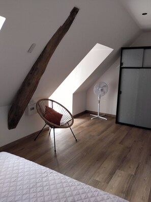 A room with a sloped ceiling, a wooden beam, a white wall, a ceiling fan, a wooden floor, and a chair with a cushion.