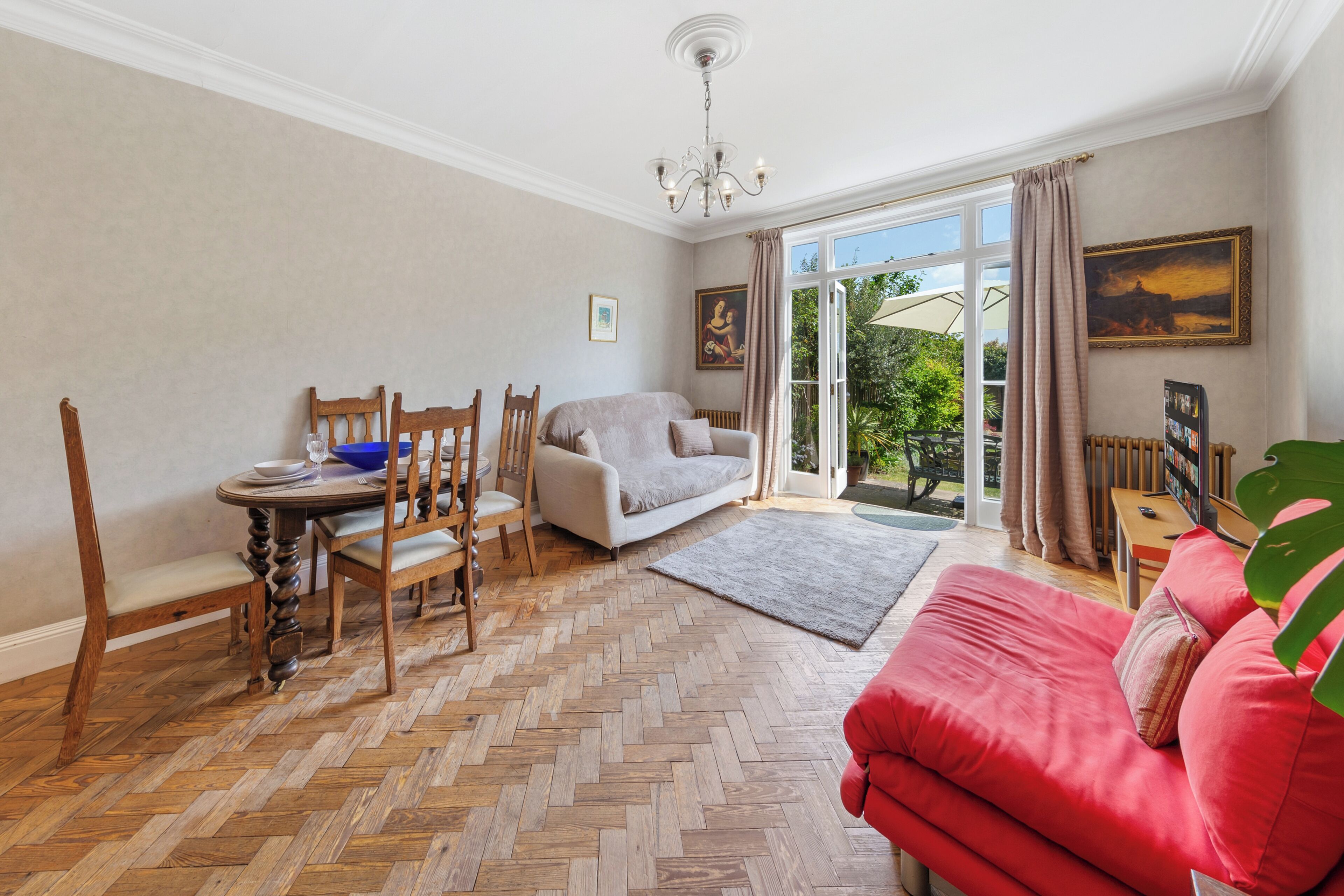 Une pièce dotée d’un parquet en chevrons, d’un canapé rouge, d’un fauteuil blanc, d’une table à manger en bois avec des chaises, et d’une grande fenêtre donnant sur un paysage verdoyant.