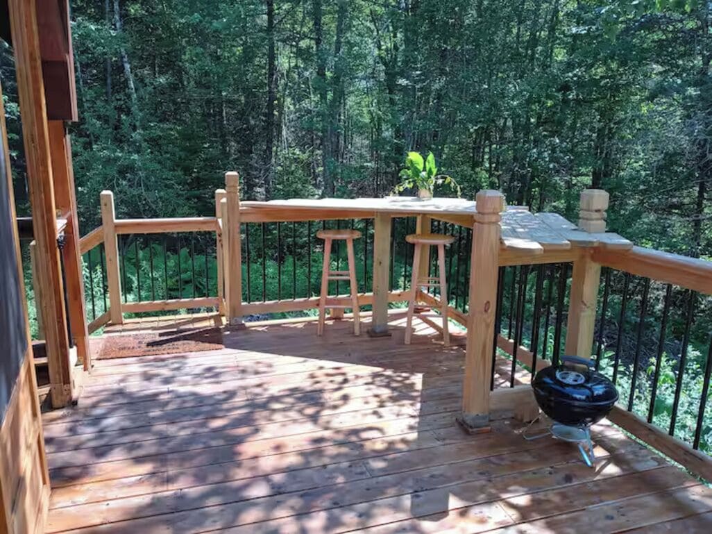 Una terraza de madera con una mesa y sillas, rodeada por una barandilla, ubicada en un área boscosa.