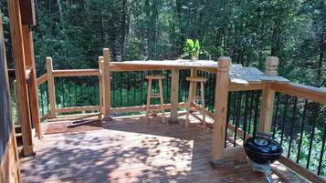 Una terraza de madera con una mesa y sillas, rodeada por una barandilla, ubicada en un área boscosa.