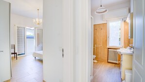 Una habitación de hotel moderna con un amplio ventanal corredero que da a un balcón, un baño independiente con puerta de madera y un candelabro.