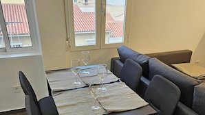 A dining area with a table set for four, a sofa, and a view of buildings through a window. - 2BR Family Flat * Balcony * Walk to Everything (Málaga)