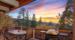 Eine Holzterrasse mit Tisch, Stühlen, Grill und Blick auf Bäume und einen Sonnenuntergang.