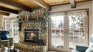 A stone fireplace with a fire, a wooden ceiling, and a view of a snowy deck through glass doors. - Hot Tub, Near Bear Mt, Lake Views, Dog Yard, AC (Big Bear Lake)