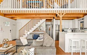 A spacious living area with a grey sofa, wooden staircase, and a kitchen with white cabinets. - Lakefront House With AC, Private Dock, Game Loft (Big Bear Lake)