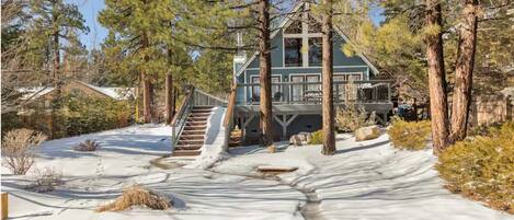 A cabin with a staircase leading to a deck, surrounded by snow and trees.