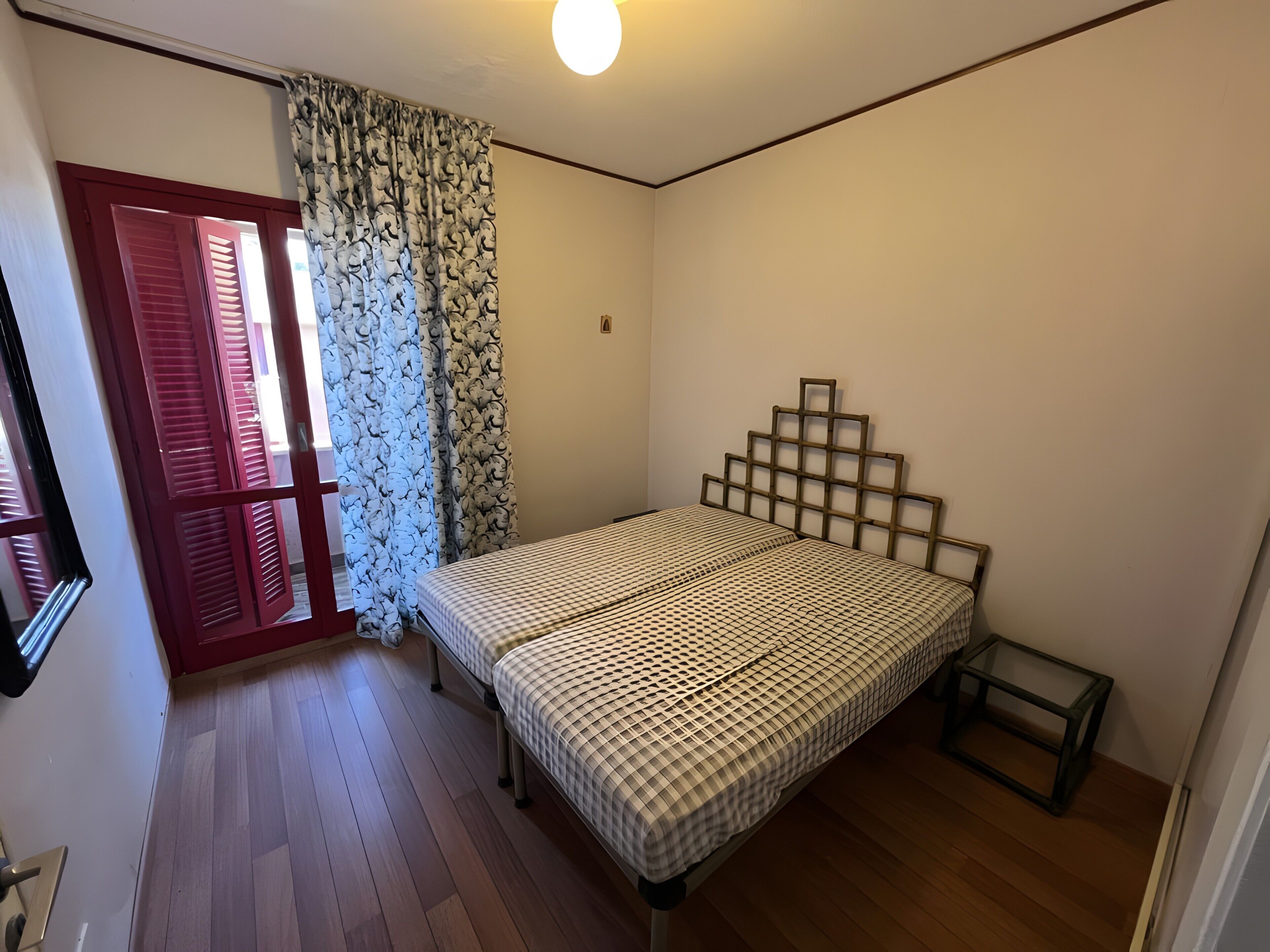 A bedroom with a bed, a small table, a curtain, and a door with shutters.