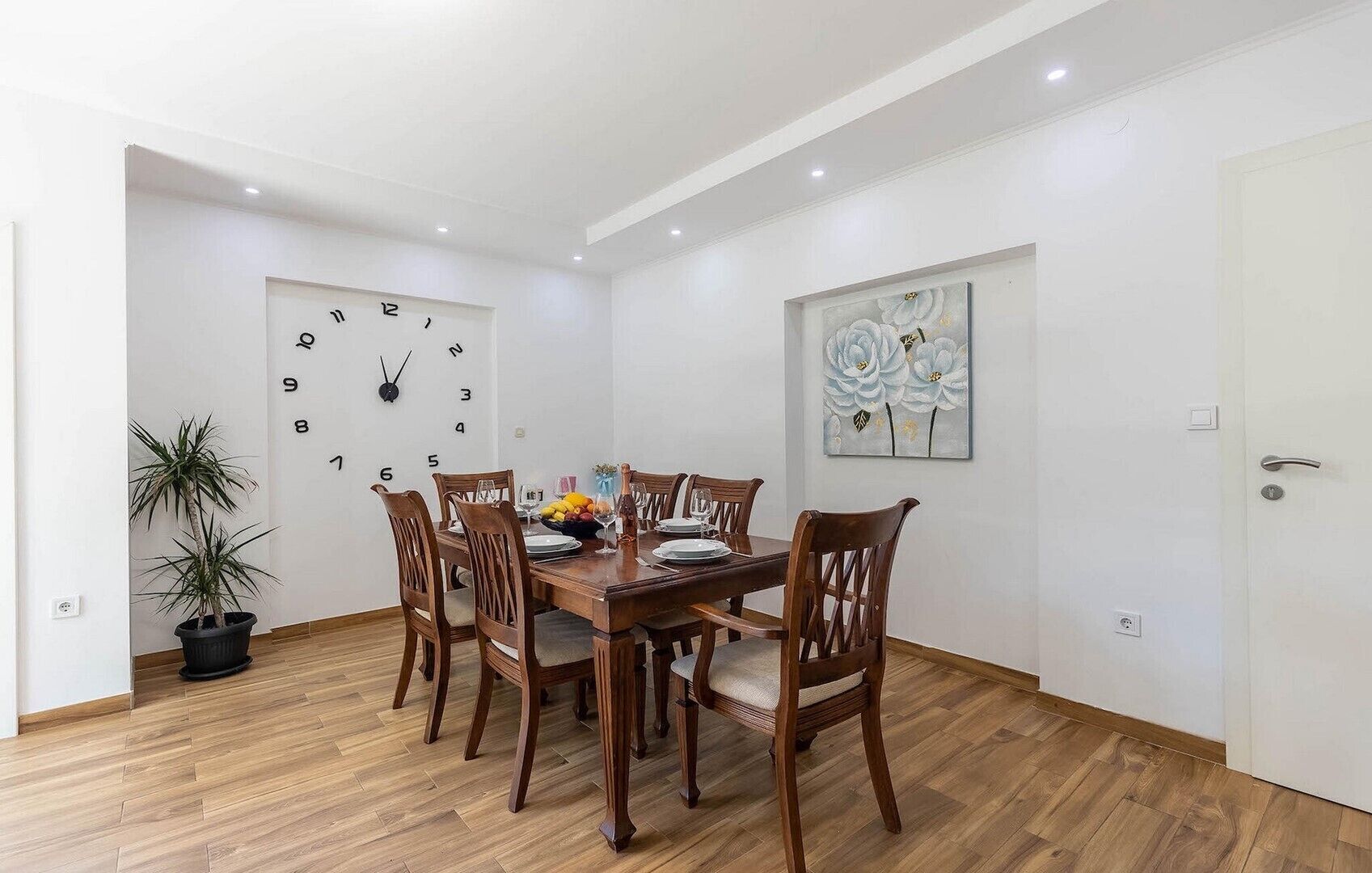 Une salle à manger avec une table en bois, des chaises, une horloge, une plante en pot et un tableau mural.