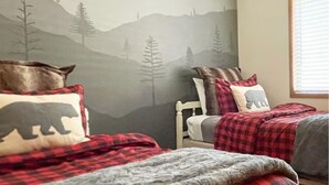 Un dormitorio con dos camas, cada una con un mural temático de montaña, una ventana con persianas y una silla con una manta a cuadros rojos y blancos.