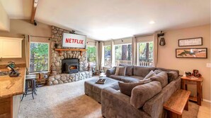 Un salon avec une cheminée en pierre, un écran affichant le logo de Netflix, un grand canapé et une vue sur les arbres à travers les fenêtres.